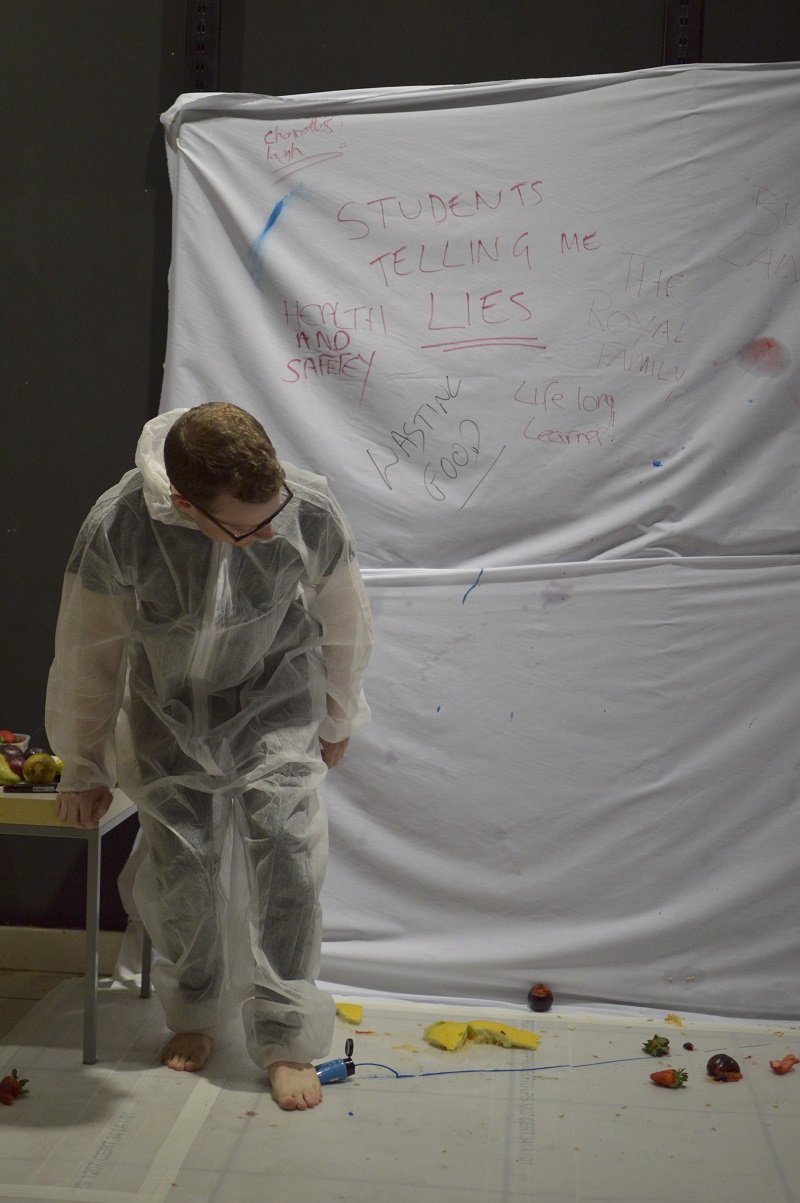 A photo of a man in a paper boiler suit, barefoot and stepping on a tube of blue paint, leaning forward in front of a canvas on which are written several reasons for being angry.